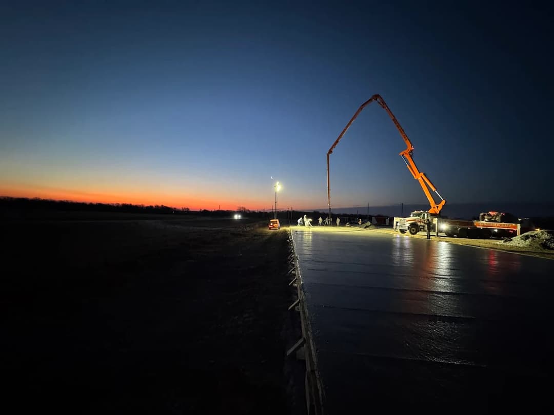 Concrete pouring at dusk with construction equipment against a colorful sunset.