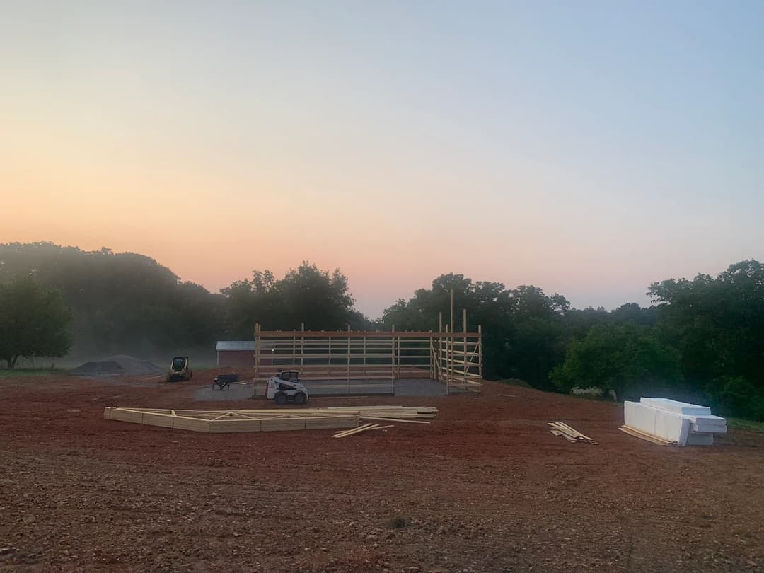 Construction site at dawn with wooden framework and equipment against a colorful sky.