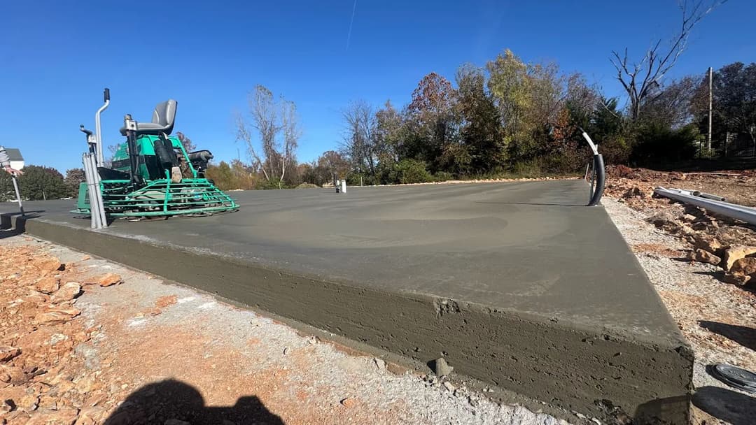 Freshly poured concrete slab with a finishing machine, surrounded by trees and clear blue sky.