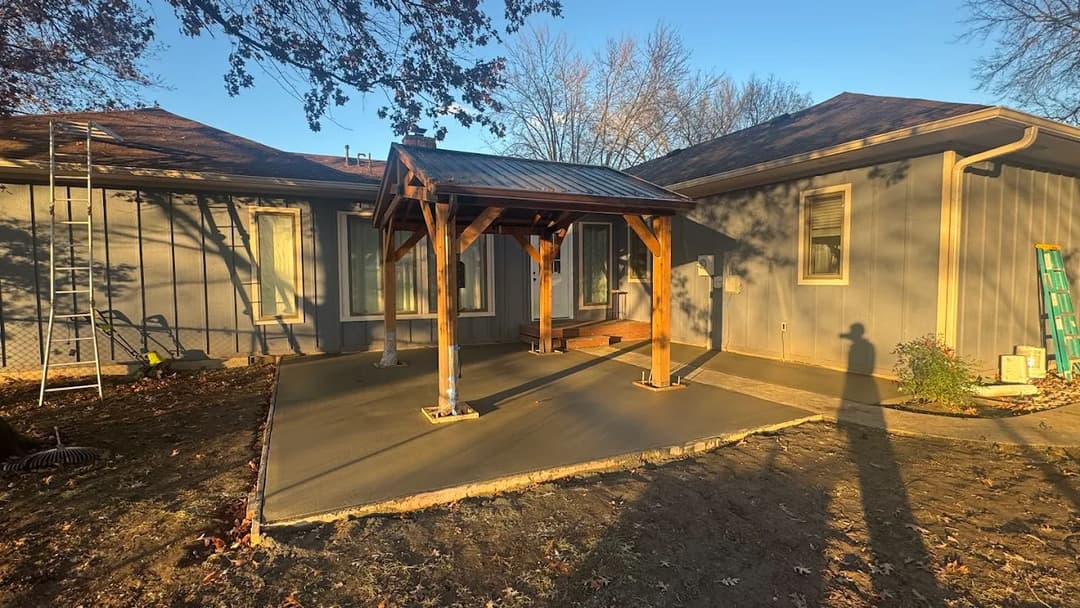 Newly poured concrete patio with wooden gazebo next to a house in a sunlit backyard.