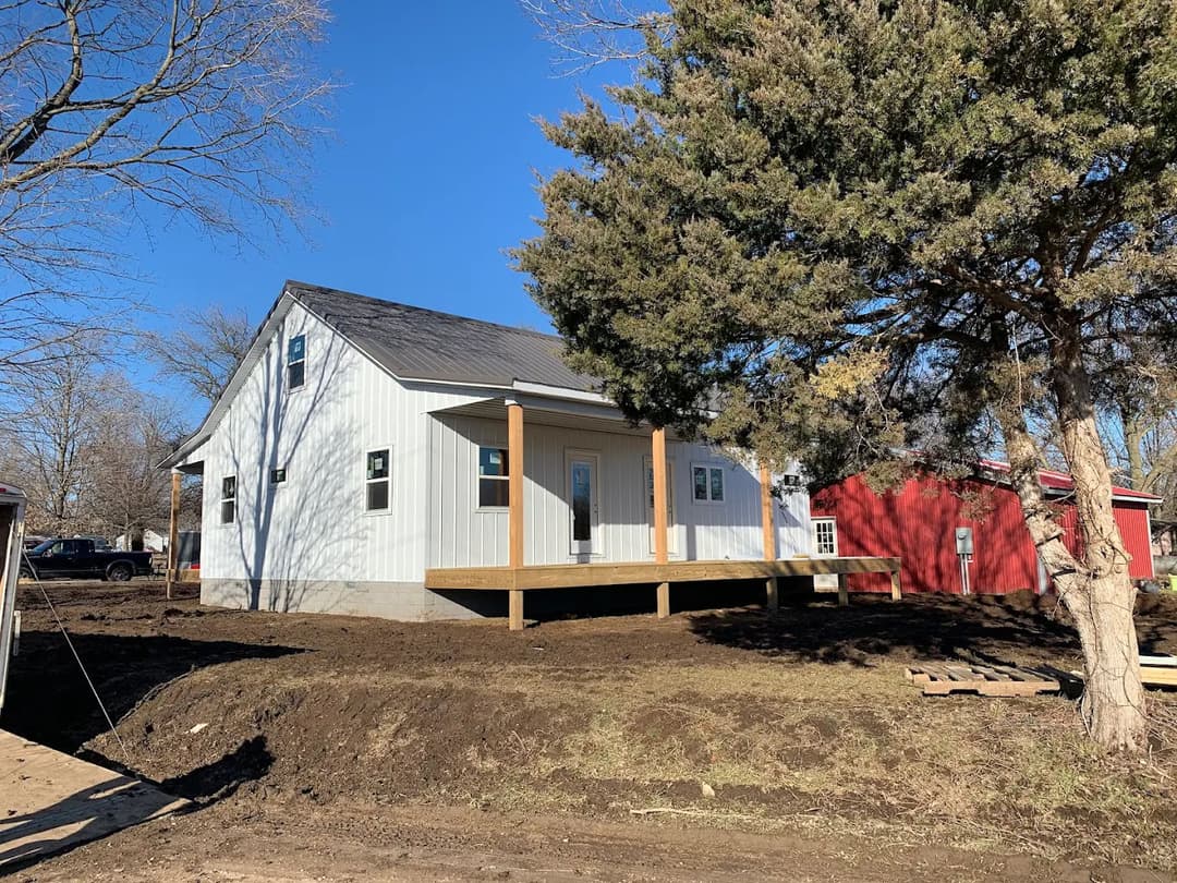 Modern white house with front porch and wooden deck, surrounded by trees and grassy area.
