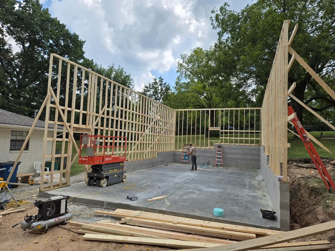 Construction site with framed walls and workers building a house. Tools and equipment visible.