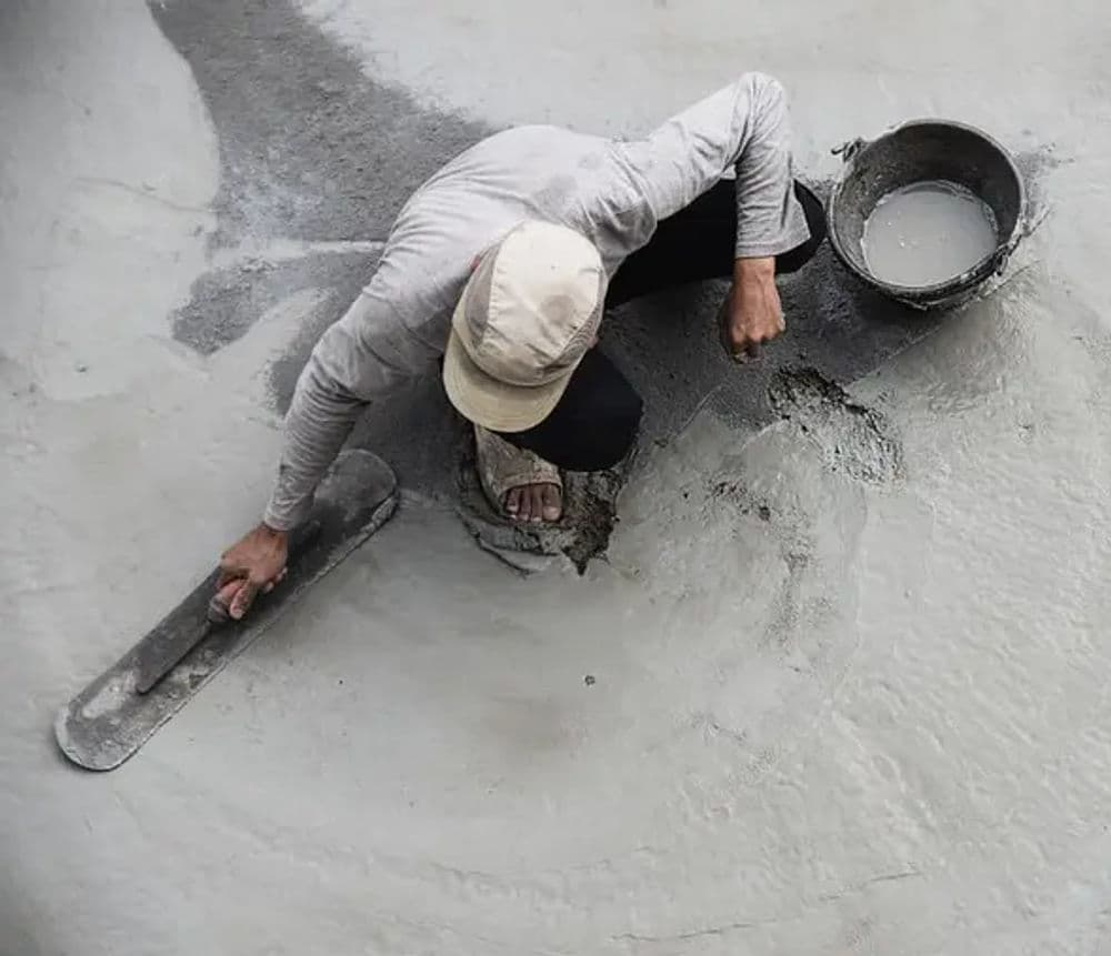 Worker smoothing wet concrete with a trowel, with a bucket of mixture nearby.